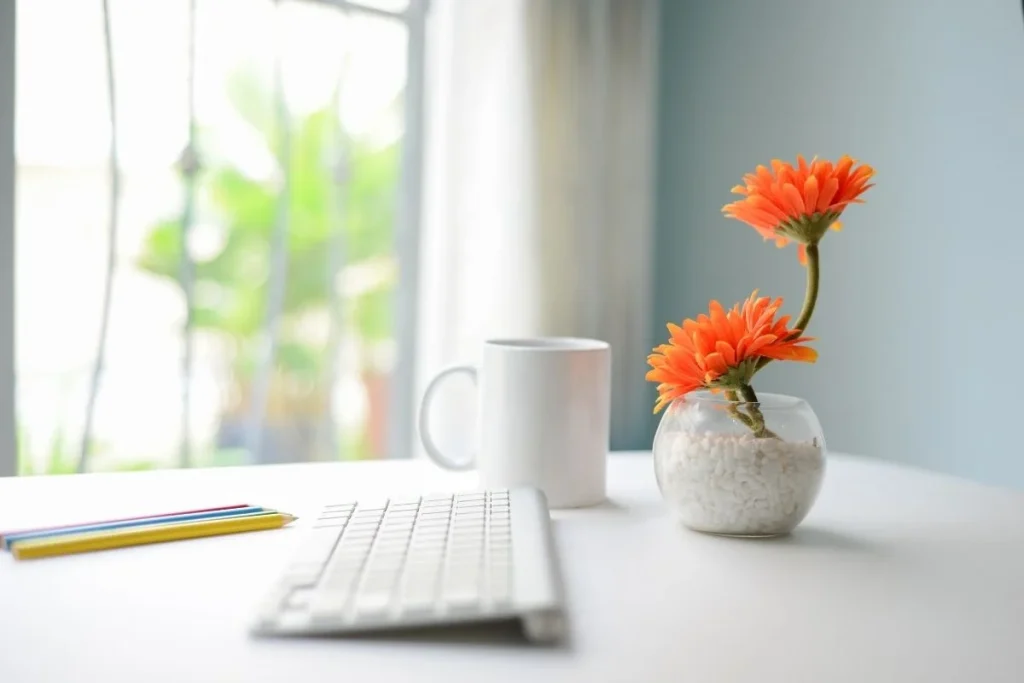 Minimalist home office desk with white keyboard, coffee mug, colorful pencils, and small glass vase holding bright orange flowers near a sunlit window.