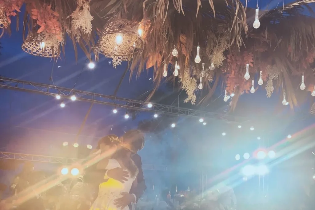 A couple sharing a warm embrace while dancing under hanging lights and floral decor at an evening celebration, symbolizing love in the ordinary moments of marriage.
